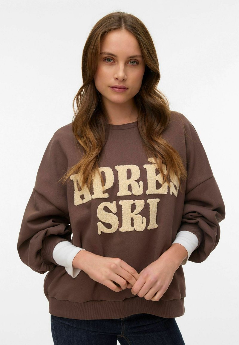 Sudadera marrón con las palabras "APRES SKI" en letras beige texturizadas. Presenta mangas anchas y un dobladillo acanalado. Se lleva con una camiseta blanca de manga larga debajo.