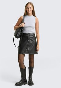 Zwarte faux leren A-lijn rok met knopen aan de voorkant, gecombineerd met een lichtgrijze mouwloze top en zwarte knielange laarzen. Schoudertas inbegrepen.