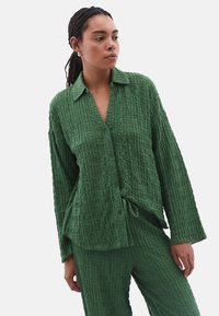 Camicia verde con bottoni, realizzata in un tessuto texturizzato e stropicciato. Presenta un colletto, maniche lunghe e una vestibilità comoda con dettaglio a laccio all'orlo.