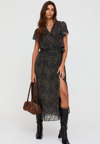 Schwarzes Maxikleid mit kurzen, gerafften Ärmeln, goldenem abstraktem Muster, seitlichem Schlitz, tailliert, kombiniert mit einer braunen Handtasche und kniehohen schwarzen Stiefeln.