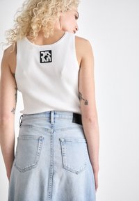 Bijela rebrasta majica bez rukava s crnim logom, u kombinaciji s svijetloplavom traper suknjom koja ima dva stražnja džepa i zatvaranje na patentni zatvarač.