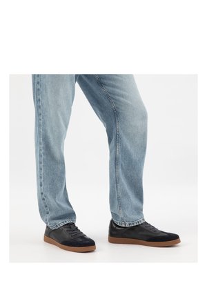 Persona che indossa jeans azzurri chiari e sneakers nere con suole marroni, in piedi su uno sfondo bianco.