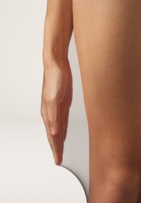 Nøgen, gennemsigtig strømpebukser der hænger over et ben, med en hånd der let rører ved stoffet. Glat tekstur og subtil glans er synlige.