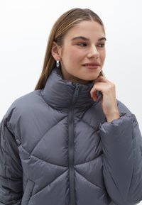 Chaqueta acolchada gris con cuello alto y cierre de cremallera. Presenta una textura suave y patrones de costura diagonal.