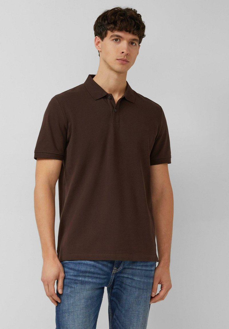 Junger Mann, der ein dunkelbraunes Poloshirt mit kurzen Ärmeln und blaue Jeans trägt, vor einem einfarbigen hellgrauen Hintergrund steht.