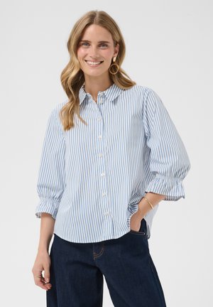 Kvinde smiler og har en lyseblå og hvidstribet skjorte med trekvart ærmer og mørkeblå jeans på, med hånden i lommen.