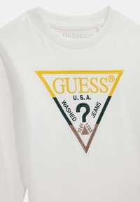 Camiseta de algodón blanca con un logotipo bordado triangular en amarillo, verde y marrón, que presenta "GUESS," "U.S.A." y "WASHED JEANS."