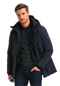 Chaqueta acolchada de color azul oscuro con capucha, que presenta un forro interior texturizado y cierre de cremallera, llevada sobre un suéter de punto oscuro y jeans de mezclilla.