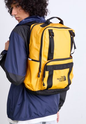 Personne portant une veste bleu marine avec un grand sac à dos jaune et noir The North Face sur le dos, tournée vers la gauche.