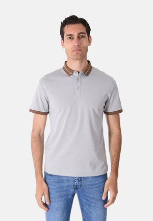 Uomo che indossa una polo grigio chiaro con colletto marrone e bordi delle maniche, abbinata a jeans blu, in piedi davanti a uno sfondo bianco.