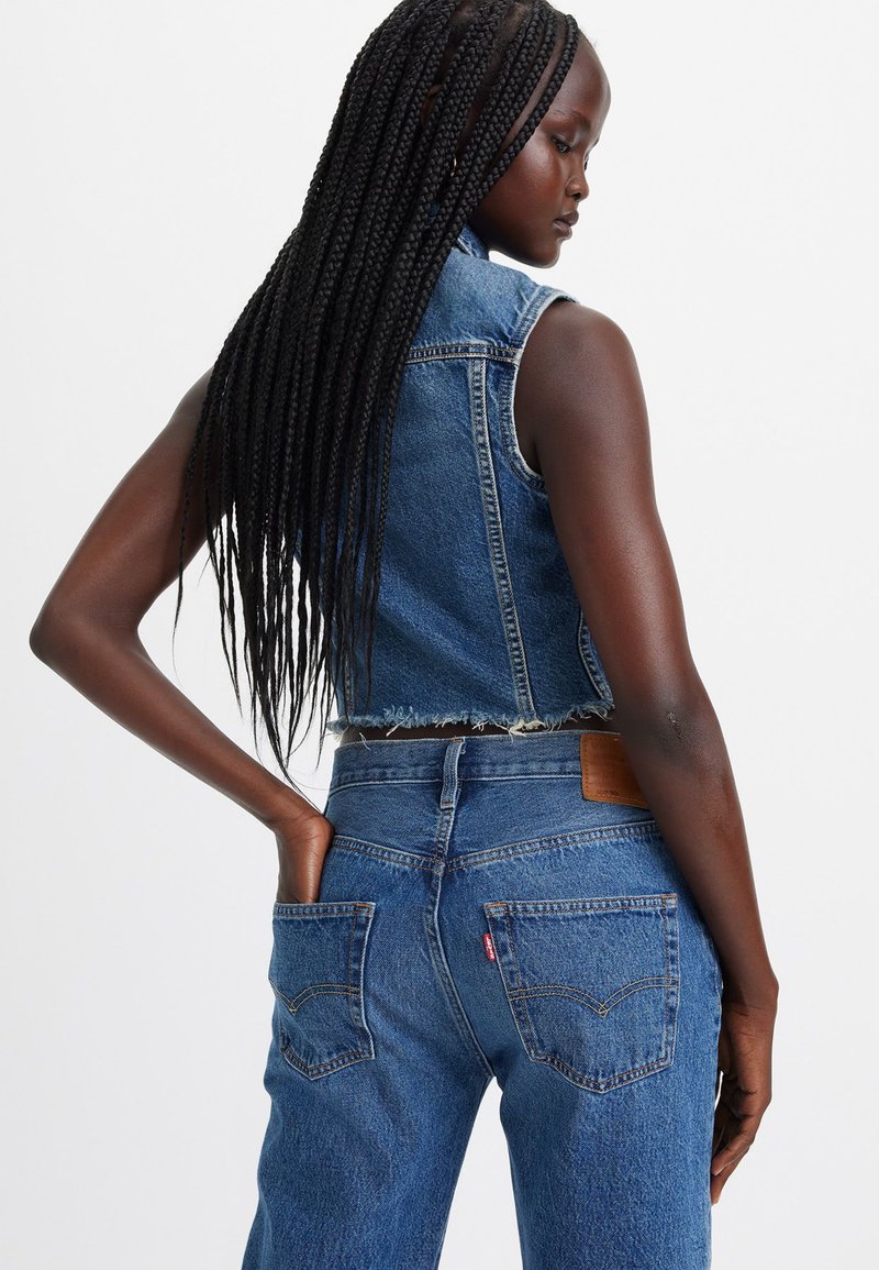 Denimweste und hochgeschnittene Jeans in mittlerem Blau. Die Weste hat ausgefranste Kanten und keine Ärmel; die Jeans verfügen über Gesäßtaschen und gestickte Details.