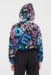Persona che indossa una giacca nera con cappuccio decorata con testo, simboli e disegni in stile graffiti colorato, abbinata a pantaloni neri.