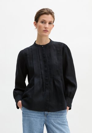 Kobieta w czarnej bluzce na guziki z pionowymi plisami i bufkami na rękawach, w jasnoniebieskich dżinsach, pozująca z rękami w kieszeniach.