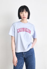 Helles blaues Baumwoll-T-Shirt mit fettem roten Schriftzug "CALVIN KLEIN" in einem gewölbten Design. Kurze Ärmel und lockere Passform.