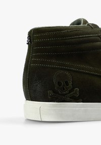 Zapatillas de ante verde oliva de caña alta con suela de goma blanca, que presentan costuras decorativas y un emblema de calavera y huesos cruzados en el lateral.