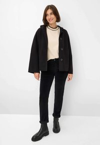 Frau mit kurzen braunen Haaren, die eine schwarze Kapuzenknopfjacke, ein cremefarbenes Oberteil, schwarze Hosen und schwarze Stiefeletten vor einem weißen Hintergrund trägt.