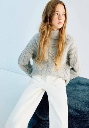 Maglione chunky lavorato a maglia grigio chiaro con collo alto e motivo a trecce, abbinato a jeans bianchi a gamba larga con dettagli in metallo.