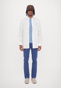 Camicia bianca a bottoni sopra una maglietta azzurra, abbinata a pantaloni blu e scarpe slip-on beige. Un cappellino marrone con logo completa l'outfit.