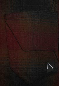 Tessuto scozzese rosso e nero con una grande tasca inclinata e cuciture nere. Un piccolo logo è visibile sull'angolo della tasca.