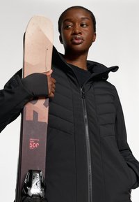 Veste zippée noire avec un devant matelassé, tenant un ski avec des accents beiges et noirs. Le ski a une prise texturée et des graphismes.