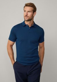 Mörkblå pikétröja med korta ärmar, ribbad struktur, tre-knappsknäppning och broderad logotyp på bröstet. Kombinerad med mörka byxor.