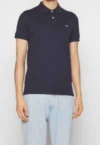 Mörkblå polo skjorta i bomull, med klassisk krage, knäppning med två knappar, korta ärmar och en liten logotyp på bröstet.