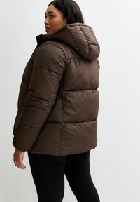 Brązowa kurtka puchowa z kapturem, z pikowanym wzorem i szerokimi poziomymi sekcjami. Czarne spodnie noszone pod spodem, minimalne tło.