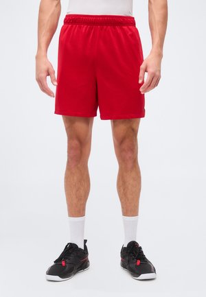 Homme portant un short de sport rouge, des chaussettes blanches montantes et des chaussures de sport noires, debout devant un fond uni.