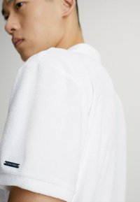 Polo shirt in terry cloth bianco con maniche corte, caratterizzato da un piccolo etichetta nera "Calvin Klein" sulla manica sinistra. Design e texture semplici.