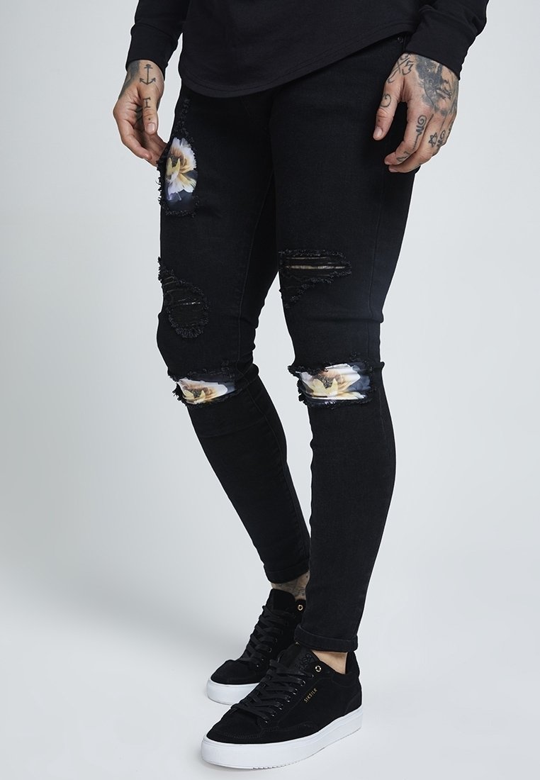 Black skinny jeans with ripped knees, revealing floral fabric underneath; paired with black sneakers featuring white soles and subtle branding.