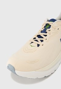 Αθλητικό παπούτσι με ελαφρύ μπεζ άνω μέρος, μπλε σκούρα κορδόνια και υφή σόλας. Περιλαμβάνει τρύπες αερισμού και λογότυπο με πράσινη λεπτομέρεια.