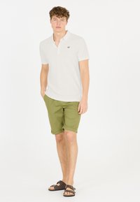 Vit pikétröja med en svart logotyp, gröna shorts och bruna sandaler. Pikétröjan har krage och korta ärmar, medan shortsen är upprullade.