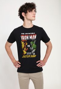 Černé tričko s komiksovým motivem Iron Mana a Doktora Dooma v červené, žluté a zelené barvě, s výrazným textem a detaily.
