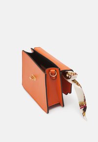 Sac à main en cuir orange avec des accents noirs, forme rectangulaire, et un détail en métal doré. Un foulard en soie à motifs est attaché sur un côté.