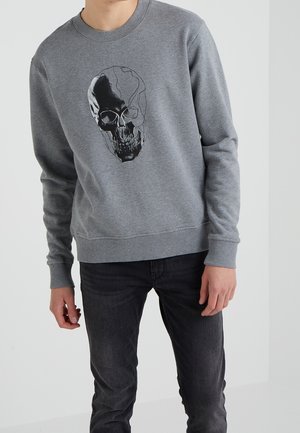 Person iført en grå sweatshirt med en sort-hvid kraniegrafik på fronten, kombineret med mørke jeans mod en ensfarvet baggrund.