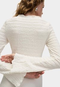 Top bianco a maniche lunghe con texture e polsini arricciati, caratterizzato da una silhouette aderente e motivi astratti. Vista posteriore che mostra i dettagli.