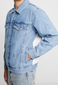 Uomo che indossa una giacca di denim blu chiaro con bottoni metallici, tasche sul petto e una striscia bianca sui lati, con una mano in tasca, tagliata all'altezza del mento.