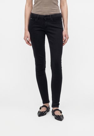 Person trägt schwarze Skinny-Jeans, ein beiges ärmelloses Oberteil und schwarze spitze Ballerinas mit silbernen Nieten und Knöchelriemen.
