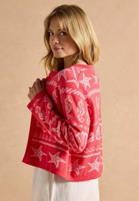 Korálově růžový vzorovaný svetr s motivy mušlí a hvězdic, dlouhými rukávy a nadměrným střihem, vystavený na neutrálním pozadí.