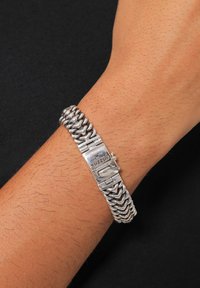 Bracciale in argento con chiusura che presenta un logo, composto da anelli intrecciati e una finitura lucida, indossato su un polso contro uno sfondo scuro.