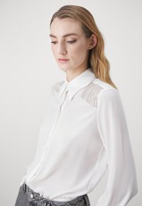 Camicia bianca a maniche lunghe con colletto, caratterizzata da dettagli in perline metalliche sulle spalle. Tessuto liscio con vestibilità sartoriale.