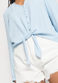 Blusa azzurro chiaro a maniche lunghe con nodo frontale, caratterizzata da piccoli bottoni e un tessuto testurizzato, abbinata a pantaloni corti di denim bianchi.