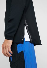 Top negro de manga larga con textura de malla, botones a presión en el lateral y una capa interior blanca de contraste, combinado con pantalones de estampado azul y negro.