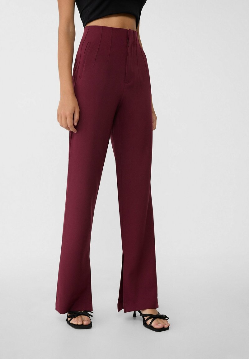 Femme portant un pantalon bordeaux taille haute à jambes larges avec des fentes à l'avant, associé à des sandales à talons noires à brides et un top court noir.