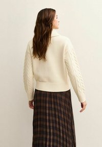 Elfenbeinfarbener Strickpullover mit einem Kabelstrickmuster an den Ärmeln und einem gerippten Saum, getragen über einem braun karierten gefalteten Rock.