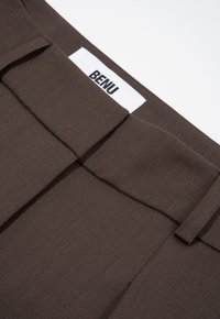 Donkerbruine stoffen broek met plooien bij de taille, met een wit label met "BENU" aan de bovenkant. Gladde textuur, op maat gemaakte design.
