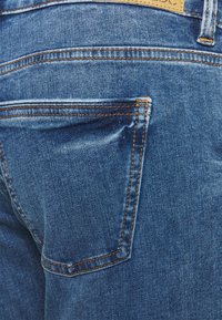 Os jeans de denim apresentam um tom azul com um acabamento ligeiramente desbotado. O bolso traseiro tem costuras de contraste laranja e uma forma retangular.