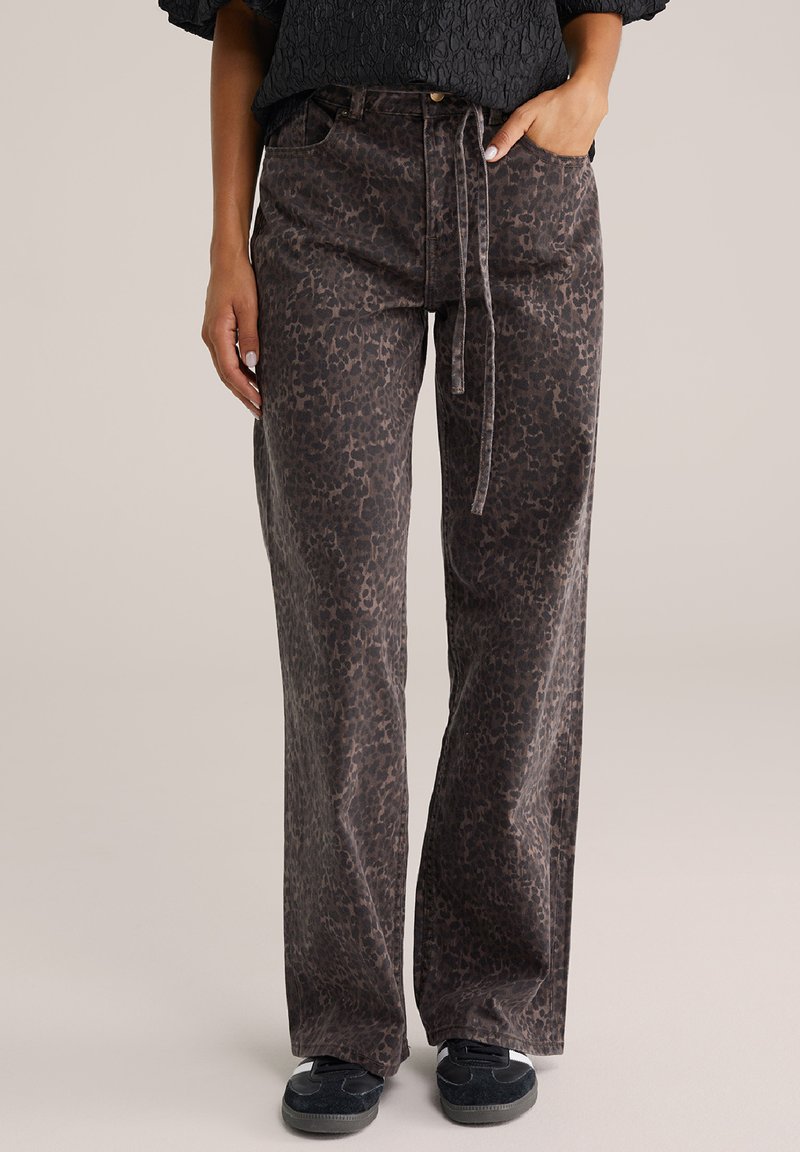 Pantalones de pierna ancha en marrón oscuro con un estampado de leopardo. Hechos de tela suave, cuentan con un cordón en la parte delantera y cinco bolsillos. Combinados con zapatos negros.