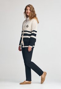 Kardigan in Creme und Marineblau mit horizontalen Streifen, ausgestattet mit einem Logo-Patch. Kombiniert mit schwarzen Hosen und lohnenden Mokassins aus beigefarbenem Wildleder.