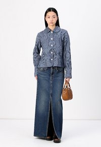 Light blue, laser-cut jacket with large buttons, paired with a long denim skirt featuring a front slit. Model holds a small brown bag.